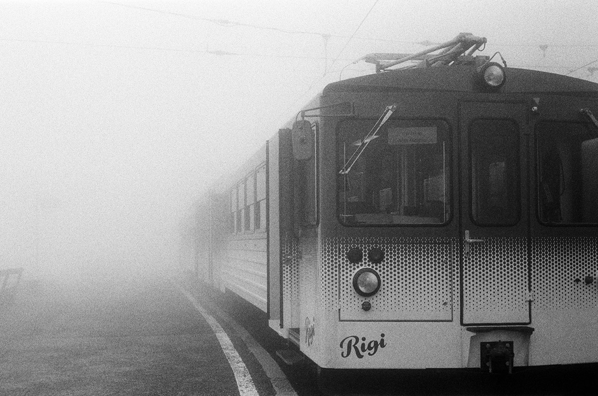 a foggy train platform with a cogwheel train labeled 'Rigi'. the fog is so thick, visibility of the train drops off after about the first car.