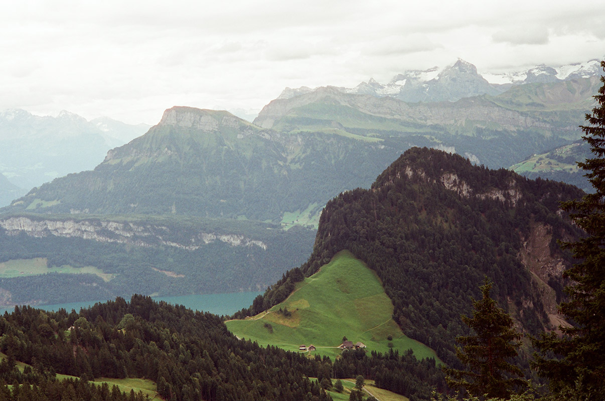 the view towards Lake Lucerne (only barely in view) looking downwards from Rigi. it's expansive with valleys in various shades of green. steep cliffs and a couple of chalets visible. in the distance, tall snowcapped mountains.