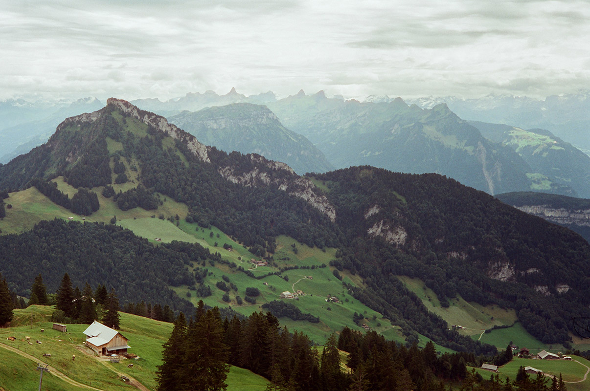 like the first picture, but without the lake in view. more farmland is visible. sharper jagged snowy alpine peaks in the distance.